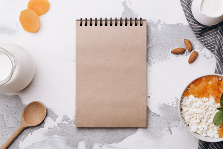 Cottage cheese with dried apricots, mint and milk on white background. Healthy breakfast concept. Empty craft notebook for recipe. Copy spaceの写真素材