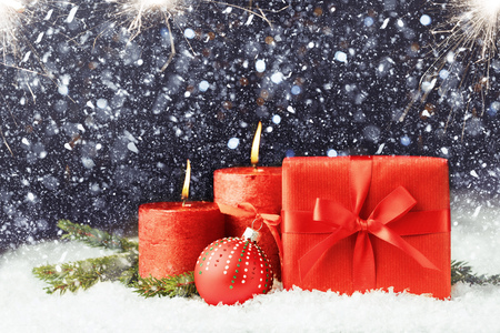 Red gift with bow on the snow in snowfall, bright and dark Christmas background with red burning candles and ball, copy spaceの写真素材