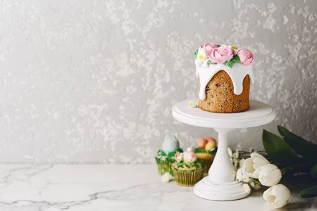 Easter cake with creamy roses on white stand. White flowers with holiday cupcakes. Cement wall. Copy spaceの写真素材
