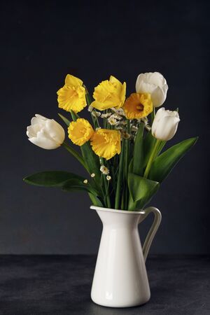 Fresh bouquet of yellow daffodils and white tulips in vase on black table. Springtime still life concept.の写真素材
