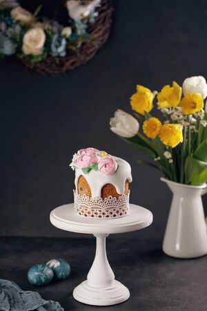 Homemade Easter cake on white wooden stand with bouquet of daffodils and tulips in vase on dark background. Holiday decoration concept.の写真素材