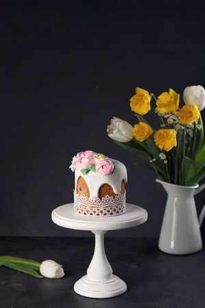 Homemade Easter cake on white wooden stand with bouquet of daffodils and tulips in vase on dark background. Holiday conceptの写真素材