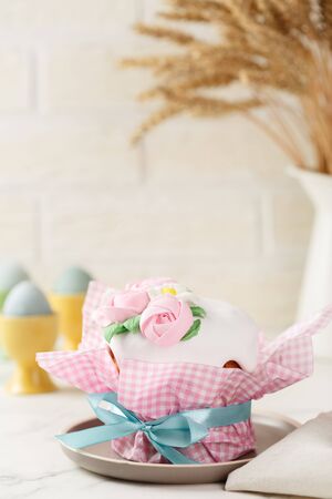 Easter cake in pink paper packaging with blue ribbon on the grey plate. Homemade festive pie with frosting and mastic floral decoration. Bunch of wheat ears at the background. Happy Easter concept. Copy spaceの写真素材