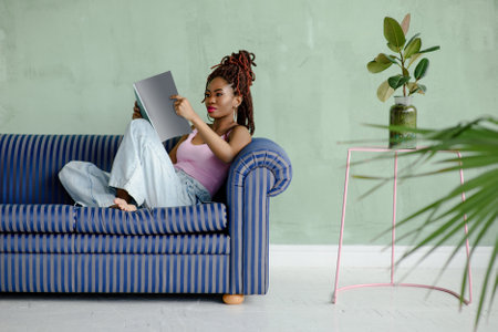 Pretty African young woman reading magazine and smiling while sitting on the sofa in a comfortable pose in a living room with light green walls and houseplants. Leisure concept.の写真素材