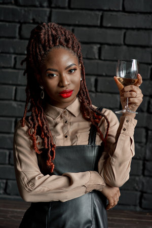 A half-body close-up portrait of a confident well-dressed African woman holding a glass of white wine against a black brick background. Wine tasting, wellness, relaxing self care. Waiting for a date.の写真素材