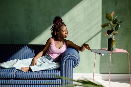 Beautiful African woman wearing casual clothes, reaching for the magazine while sitting on the sofa...