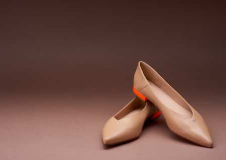 Fashionable ballerina shoes with pointed toes and low bright orange heels isolated on a gradient brown background. One shoe stands on the other. Copy space for text. Studio shot of trendy footwearの写真素材