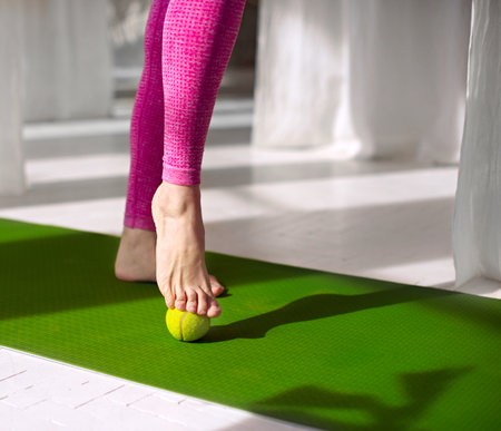 Massage for feet with tennis ball. Woman practicing foot self-massage with a tennis ball. Physiotherapy, reflexology. Tension relieving in the plantar fascia. Improving foot joint mobility.の写真素材