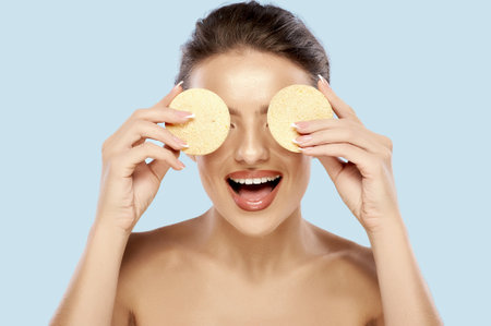 A cheerful young woman with french manicure holds two cellulose sponges in front of her eyes isolated over a gray background. Makeup removing. Creative layout for facial cleansing product ads.の写真素材