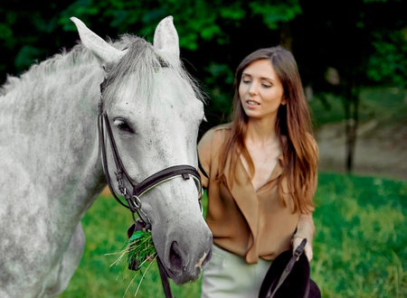 Young girl standing with a grey horse on a grass. Learn horseback riding through lessons. Explore horse therapy, hippotherapy. A woman and her grey horse in a forest, bonding withの写真素材