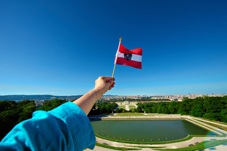 Close-up of a woman's hand holding an Austrian flag against imperial Wien SchÃ¶nbrunn Palace with Vienna city panorama in the background, captured from the Gloriette cafe roof on a summer afternoon.の写真素材