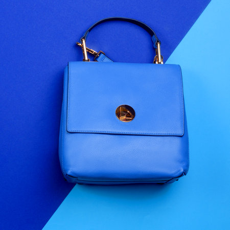 Stylish female blue leather flap bag with top handle, and gold hardware on a graphic two-tone blue background. Discount, season sales. Retail, store, boutique. Fashion blog or magazine concept.の写真素材