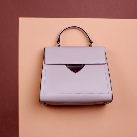 Fashionable beige leather female top-handle bag with black trim, metal accent, and feet isolated on a two-tone graphic background with copy space. Creative mockup for advertising a handbag store.の写真素材