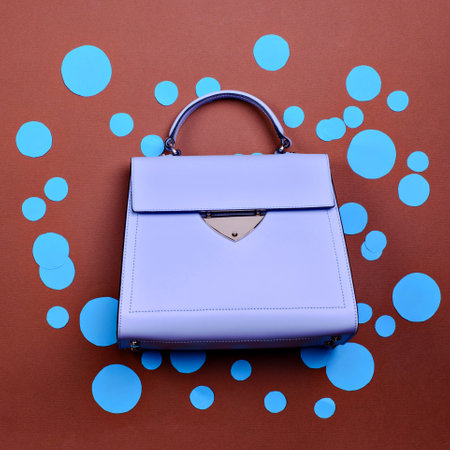 Trendy lavender leather female top-handle bag with black trim, metal accent, and feet isolated on a cartoonish brown paper background with blue circles or bubbles. Creative artwork. E-commerce store.の写真素材