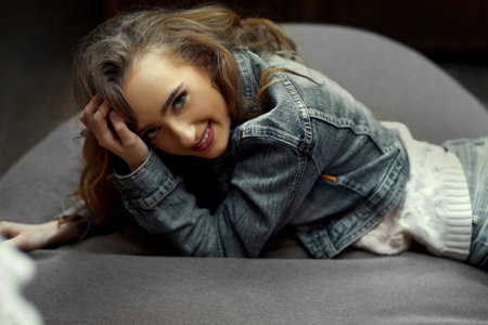 Young pretty girl with cherry lipstick dressed in a denim jacket with a white sweater is smiling while lying on a gray beanbag in the living room. Inner peace, calmness and relaxation conceptの写真素材