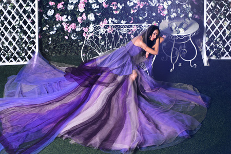 Beautiful brunette woman wearing long purple dress sitting on the bench in a flower gardenの写真素材