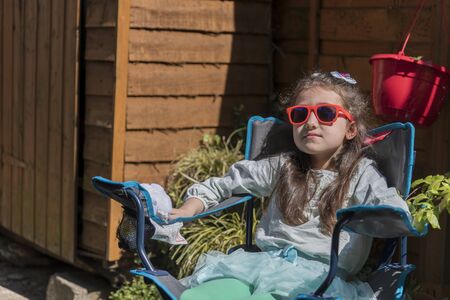 Young caucasian girl with a sad face holding a toy, sitting on bench in the yard. Portrait of child with a toy outdoors.の写真素材