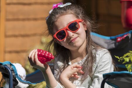 Young caucasian girl with a sad face holding a toy, sitting on bench in the yard. Portrait of child with a toy outdoors.の写真素材