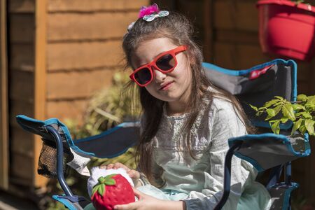 Young caucasian girl with a sad face holding a toy, sitting on bench in the yard. Portrait of child with a toy outdoors.の写真素材