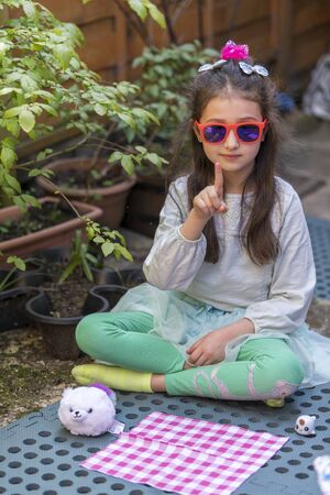 Young caucasian girl with a sad face holding a toy, sitting on bench in the yard. Portrait of child with a toy outdoors.の写真素材