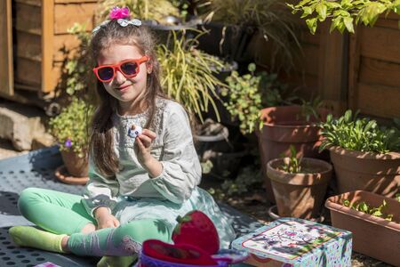 Young caucasian girl with a sad face holding a toy, sitting on bench in the yard. Portrait of child with a toy outdoors.の写真素材