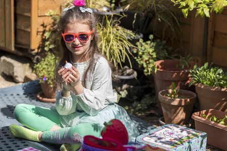 Young caucasian girl with a sad face holding a toy, sitting on bench in the yard. Portrait of child with a toy outdoors.の写真素材