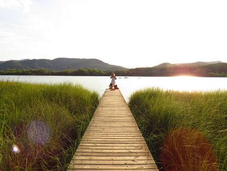 Banyoles, Spain, August 14, 2019: Footbridge on the loch Banyoles. Backlit image Silhouette of mother and two girls on their backs. Drawing a heart with the hand. Sunset over a lake. Mountains in the backgroundの写真素材