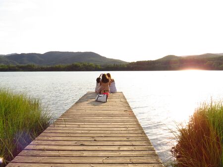 Banyoles, Spain, August 14, 2019: Footbridge on the loch Banyoles. Backlit image Silhouette of mother and two girls on their backs. Drawing a heart with the hand. Sunset over a lake. Mountains in the backgrounの写真素材