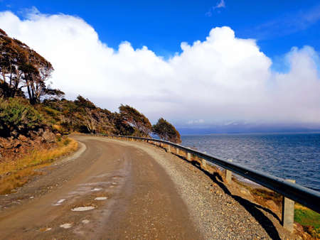 Panoramic view of road in sharp curve, trees overcome by the wind, cloudy sky and gray sea of Tierra de Fuego Argentina. Landscapes and extreme nature.の写真素材