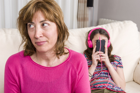Mother are angry with her daughter. Teen girl with pink headphones is listening music with her phone at home.の写真素材