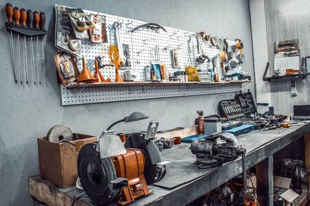 Garage. Workspace moto auto workshop closeup. Workbench with vise, grinder, sets of keys, tools kit,  equipment, spare parts. Old tools hanging on steel wall in the workshop as background, Tool shelfの写真素材