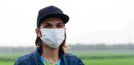 Handsome young man with long hair in white regular cotton surgical, medical mask on green forest background. Coronavirus, covid-19 protection. Banner for web site. Portrait made in front closeupの写真素材