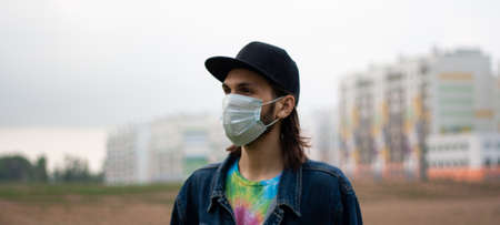 Handsome young man in cap and regular disposable surgical, medical mask on the street in city in...