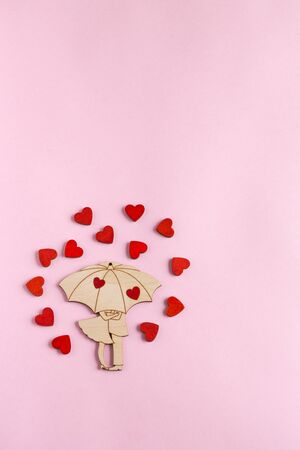 Valentine's day. A wooden figure in the form of a couple under an umbrella is located on a paper pink background. Around the pair are small, wooden, red hearts.の写真素材