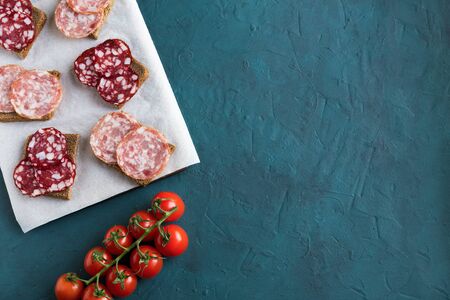 Sandwiches with smoked salchichon and braunschweig sausages are on a paper on the board. Next to are cherry tomatoes on a green background. Flat lay, horizontal composition is on a green background.の写真素材