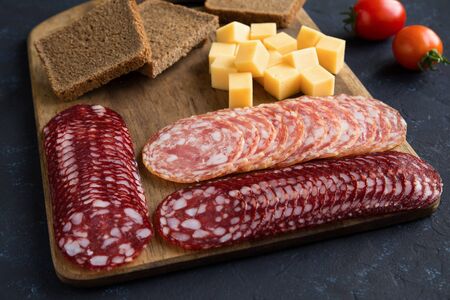 Serving of the slices of smoked salchichon and braunschweig sausages in several rows are on wooden boards. Next to it is a cherry tomato and cheese cubes. Flat lay, horizontal composition is on a dark blue background.の写真素材