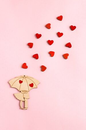 Valentine's day. A wooden figure in the form of a couple under an umbrella is located on a paper pink background. Around the pair are small, wooden, red hearts. Flat lay composition with copy space.の写真素材
