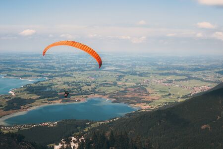 Paraglider flies from Mount Tegelbergbahn on an orange paraglider over a beautiful landscape with lakes Bannwaldsee and Forgenseeの写真素材