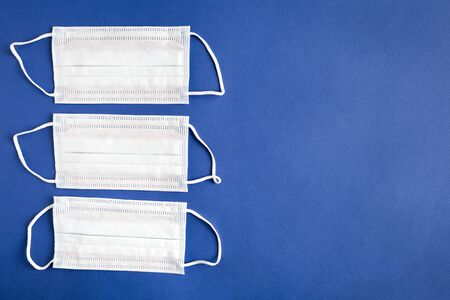 Three blue medical masks lie on a blue paper background. Medical device for breathing and protection against pollution, virus, flu and coronavirus. Flat lay on blue background with copy space.の写真素材