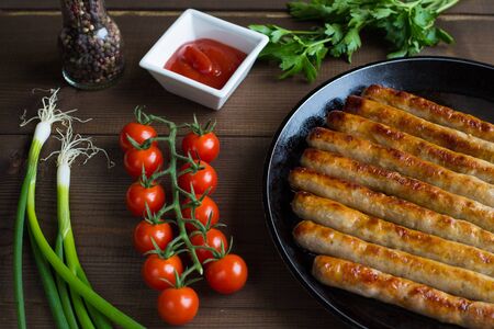 Grilled, whole, delicious sausages lie on the frying pan, which stands on a wooden background of boards. Nearby are a branch of fresh cherry tomatoes, onion feathers, parsley and ketchup in a gravy boat.の写真素材