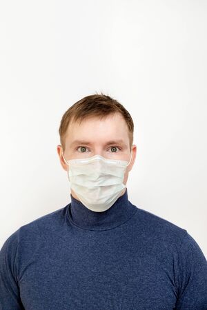 Portrait of a young man in a dark sweater on a white isolated background with copy space. He is wearing a protective mask that protects against infection, viruses, and coronavirus. His eyes are scaredの写真素材