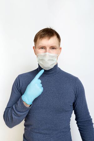 Young man wearing blue medical rubber gloves points at his face. He has medical mask on his face that protects from viruses, infection, pollution and coronavirus. Portrait on white isolated backgroundの写真素材
