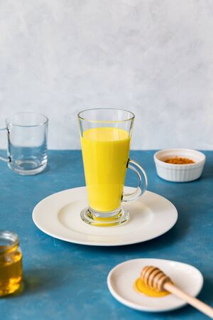 A tall glass with a herbal, delicious, traditional Indian drink with turmeric stands on a white flat plate. Golden milk for disease prevention and immune support. Next to it are the ingredients curcuma and honey. Vertical composition with copy spaceの写真素材