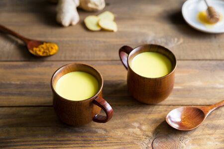 Herbal, healthy, Indian drink golden milk with turmeric root powder immune booster in two wooden cups stand on a wooden background. Next to it are two wooden spoons and the ingredients honey, ginger and curcuma.の写真素材