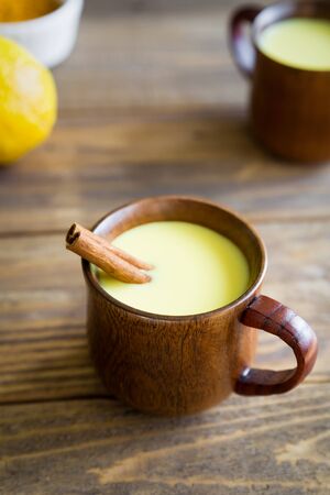 Traditional Indian drink with turmeric root powder in a wooden cup with a cinnamon stick on a wooden background. Delicious, hot Golden milk to support the immune system and prevent diseases. Herbal medicine an anti-inflammatory.の写真素材