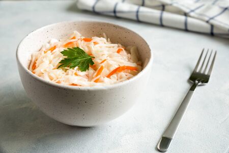 Homemade sauerkraut in a white ceramic bowl. A metal fork and a napkin lie nearby. The dish is decorated with a parsley leaf. Fermented food. Composition close up on a white background.の写真素材
