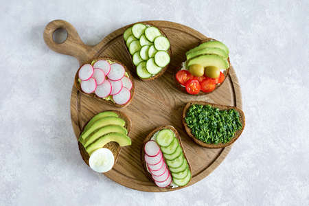 Vegetarian sandwiches on the wood board on the gray background. Fresh bread with pesto, cucumber, avocado, cherry tomatoes, radish and cheese. Flat lay, top view.の写真素材