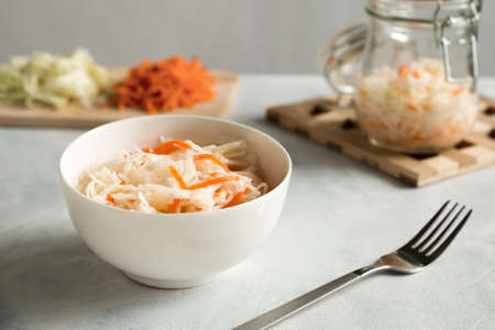 White ceramic bowl with homemade appetizer sauerkraut on a gray, concrete background. Glass jar and ingridients chopped coleslaw and carrot. Close up composition.の写真素材
