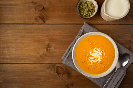 Homemade pumpkin soup in a white plate with cream and seeds on gray napkin flat lay on brown wooden rustic background with copy space.の写真素材