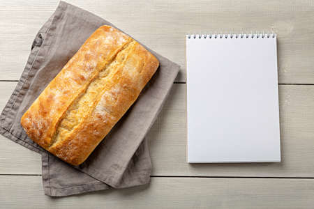 Loaf of fresh whole bread on gray napkin with notepad for recipes top view on white wooden background with copy space.の写真素材
